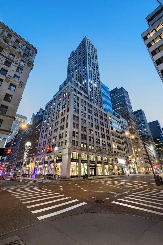The stone tower of 580 Fifth Avenue rises above its arched shopfronts at the corner of West 47th Street at dusk, with lit windows and an empty crosswalk.