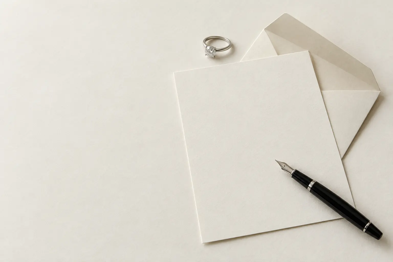 A blank sheet of cream letter paper lies on a desk with a black fountain pen, an open envelope and a diamond solitaire ring.